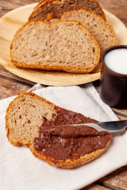 Bıçak, bir dilim kepekli ekmeğe çikolata kreması sürüyor. Arka planda daha fazla dilimler ve bir fincan süt var. Sağlıklı ve lezzetli bir kahvaltı öneriyor.