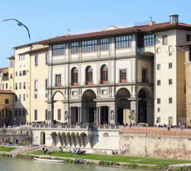 Floransa 'daki Uffizi Galerisi yakınlarındaki Arno nehri kıyısında güneşli bir günün tadını çıkaran turistler, canlı şehir manzarasının üzerindeki açık mavi gökyüzünde yükselen bir martı ile