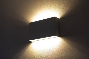 Modern duvar lambası sıcak, davetkar bir ışık yayıyor, loş aydınlatılmış bir odada duvarın dokulu yüzeyine yumuşak bir parıltı saçıyor, rahat ve atmosferik bir ortam yaratıyor.