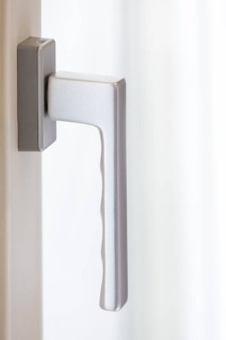 Beyaz bir çerçeveye bağlı parlak, gümüş bir pencere kulpu yakın bir pencereden yumuşak, yayılmış ışığı yakalayıp yansıtarak, temiz ve modern bir estetik yaratıyor.