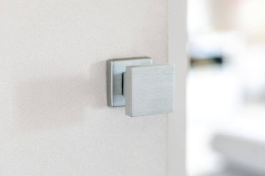 Beyaz bir kapıya monte edilmiş, modern kare fırçalı metal kapı kolu, bulanık bir iç mekan ortaya çıkarıyor, minimalist tasarım ve çağdaş ev dekorunu vurguluyor.