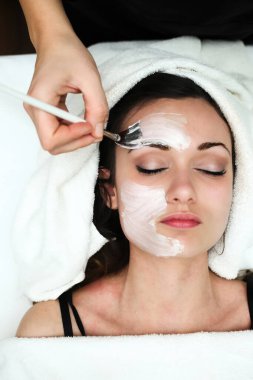 Güzellik uzmanının eli, bir fırçayla masaj yatağında gözleri kapalı, bir sağlık merkezinde yatan genç bir kadının yüzüne beyaz bir yüz maskesi takıyor.