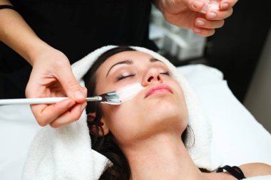 Güzellik uzmanı beyaz nemlendirici yüz maskesi uyguluyor genç esmer bir kadının yüzüne, beyaz bir yatakta bir fırçayla yatıyor, diğeri ise küçük şeffaf bir kap tutuyor.