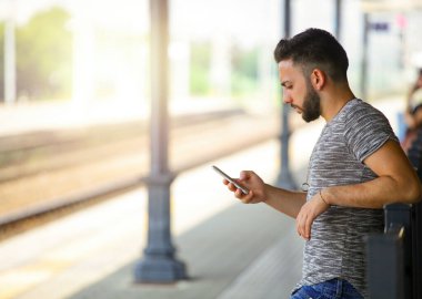 Parlak, güneşli bir günde istasyonda tren beklerken akıllı telefonunu bağlı kalmak için korkuluklara yaslanan sıradan giyinmiş genç bir adam.