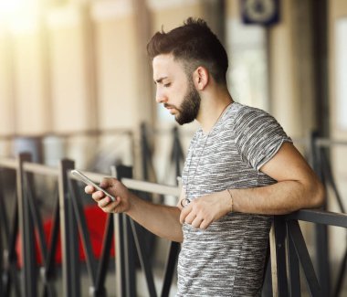 Genç sakallı bir adam tren istasyonunda korkuluklara yaslanıyor, güneşli bir yaz gününde ulaşımı beklerken akıllı telefon kullanıyor, canlı atmosferin tadını çıkarıyor.