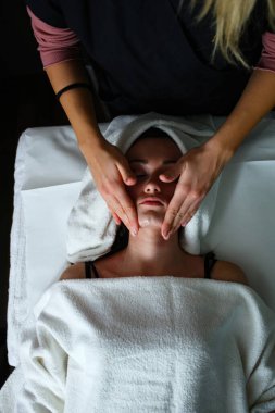 Profesyonel güzellik uzmanı masaj salonundaki ya da spa salonundaki genç bir bayana nazikçe krem sürüyor ve rahatlatıcı bir yüz masajı yapıyor.