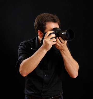 Siyah tişört giyen erkek fotoğrafçı, profesyonel bir dijital kamerayla karanlık bir arkaplan karşısında fotoğraf çekmeye odaklanıyor, profesyonelliği ve fotoğrafçılığı somutlaştırıyor.