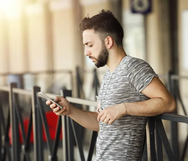 Genç sakallı bir adam tren istasyonunda korkuluklara yaslanıyor, güneşli bir yaz gününde ulaşımı beklerken akıllı telefon kullanıyor, canlı atmosferin tadını çıkarıyor.