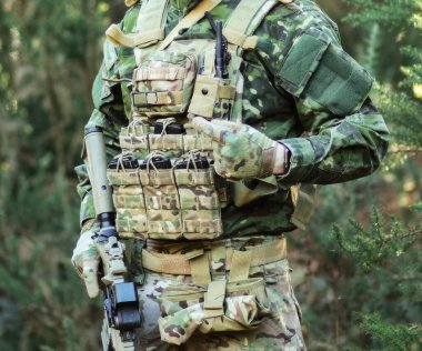 Kamuflaj üniformalı asker ve taktik yelek. Ormanlık alanda savaş eğitimi sırasında cephane dergilerine bakıyor. Savaşa hazır olduğunu ve hazırlandığını gösteriyor.