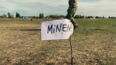 Askeri üniformalı bir adam Minen yazıtlarının arka planında mayınlı bir alanda yürüyor. Bölgenin istihkamcılar tarafından boşaltılması.