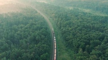 Drone tracking shot of Cargo train, freight transportation logistics concept. Freight Cargo Train with wagons shipping goods. Aerial drone shot over beautiful picturesque green foggy forest.