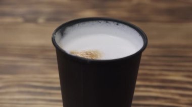 Biodegradable black paper cup with hot coffee and milk foam on the table. Coffee drink in a black glass. Coffee with milk and milk foam in a black paper cup on a wooden table.