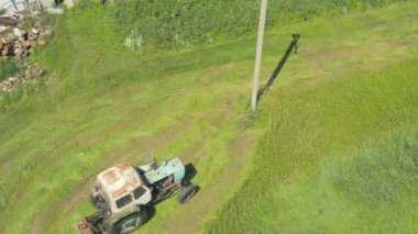 Yaşlı traktör beton bir direğin etrafındaki çimleri dikkatlice biçer. Summer Haymaking traktör çim biçme makinesiyle. Traktör saman kurutmak için yeşil çim biçer - üst hava görüntüsü. 