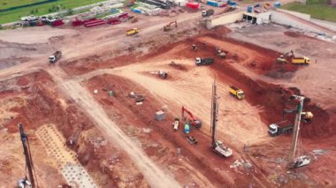 Ağaçlar, çimenler ve şehir manzarasıyla çevrili çeşitli makinelerle çevrili bir inşaat alanının havadan görüntüsü. Ekipman ve odun yığınları arasında binalar, yollar ve asfalt görülebilir