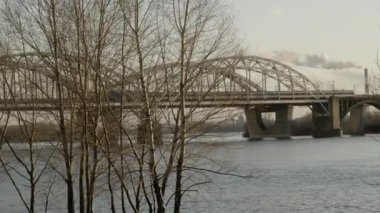 Nehir, ağaçlar ve dumanlı bir köprünün yanında gezinen bir çifti gösteren sakin bir video. Bulutlu bir gökyüzünün altında yemyeşil bir tren geçiyor.