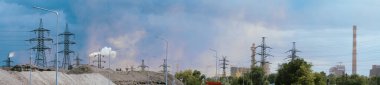 Panoramic shot of industrial area featuring power lines, smoke stacks, and a mix of natural and industrial elements under a partly cloudy sky. Juxtaposition of industry and environment.