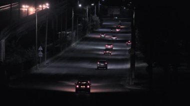 Sakin bir gece sahnesinde, sokak lambaları gece sürüşünün deneyimini artıran sakinleştirici bir atmosfer yaratırken boş bir şehir yolunda ilerleyen aydınlık araçlar görülüyor.
