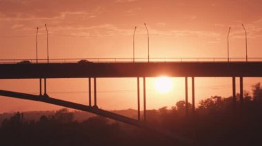 Güneş batarken, gökyüzünde ılık turuncu ve pembe renkler bırakır, aşağıdaki köprüyü siluete geçirir, çarpıcı fotoğrafçılık için mükemmel bir atmosfer yaratır.