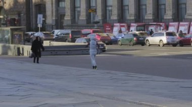 Şık, gri bir kıyafet giymiş bir şahıs, arka planda ikonik binalar tezgahlanırken, hareketli ve sayısız park halindeki arabalarla dolu bir şehir meydanında rahatça yürüyor.