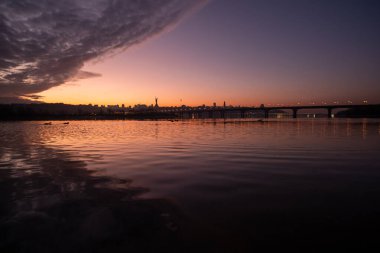 Gündüz geceye geçerken, çarpıcı bir gün batımı gökyüzünü canlı renklerle boyuyor, sakin bir nehri parlaklaştırıyor ve şehrin ufkunu aydınlatıyor, takdir etmek için büyüleyici bir manzara yaratıyor.