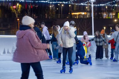 KYIV, UKRAINE - 26 Ocak 2025: Farklı yaşlardaki insanlar, festival ışıklarıyla aydınlanan Kyiv 'de açık hava buz pateni pistinin tadını çıkarıyorlar. Beyaz ceketli bir kadın paten kayarken, diğerleri buzda konuşup pratik yapıyor.