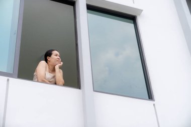 Neşeli bir kadın ikinci kat penceresinden dışarı bakarken temiz havayı soluyor, anı kucaklıyor.