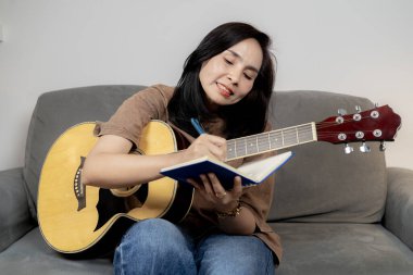 Evde akustik gitar çalarken koltukta oturup müziğin tadını çıkaran rahat bir kadın..