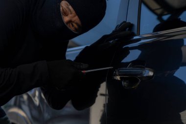 Siyah elbiseli bir suçlu gece park halindeki bir arabaya girmeye teşebbüs, araba hırsızlığı, güvenlik riski ve suç önleme kavramı.