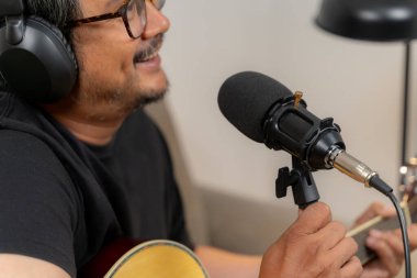 Genç adam aynı anda gitar çalıyor ve evde bir podcast kaydediyor, yaratıcılık, dijital içerik oluşturma, müzik ve modern yaşam tarzını gösteriyor