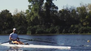 Tek başına kürek çek. Genç beyaz erkek kürekçinin yan görüntüsü, bir kürek antrenmanı sırasında, Dinyeper nehrinde bir botta oturan sporcu, sonbaharda sakin bir suda kürek çekiyor. 4K görüntü. Beyaz bir adam bir kayık eğitiyor. Ukrayna, Kyiv 'deki şehir alanı.