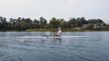 Tek başına kürek çek. Genç beyaz erkek kürekçinin yan görüntüsü, bir kürek antrenmanı sırasında, Dinyeper nehrinde bir botta oturan sporcu, sonbaharda sakin bir suda kürek çekiyor. 4K görüntü. Beyaz bir adam bir kayık eğitiyor. Ukrayna, Kyiv 'deki şehir alanı.