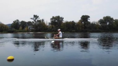 Tek başına kürek çek. Genç beyaz erkek kürekçinin yan görüntüsü, bir kürek antrenmanı sırasında, Dinyeper nehrinde bir botta oturan sporcu, sonbaharda sakin bir suda kürek çekiyor. 4K görüntü. Beyaz bir adam bir kayık eğitiyor. Ukrayna, Kyiv 'deki şehir alanı.