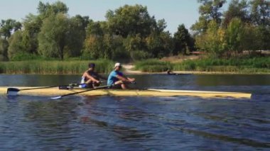 Kürek takımı eğitimi. İki beyaz erkek kürekçinin yan görüntüsü, bir kürek antrenmanı sırasında, Dnipro nehrinde bir sandalda oturan sporcu, sonbaharda sakin suda kürek çekiyor. 4K görüntü. Ukrayna, Kyiv 'deki şehir alanı.