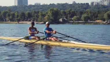 Rowing team training. Side view of 2 young caucasian male rowers, during a rowing practice, athlete sitting in a boat in the river Dnipro, rows through a calm water in autumn. 4k footage. City area in Kyiv, Ukraine.