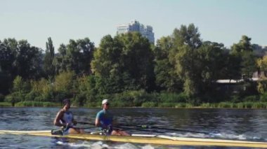Kürek takımı eğitimi. İki beyaz erkek kürekçinin yan görüntüsü, bir kürek antrenmanı sırasında, Dnipro nehrinde bir sandalda oturan sporcu, sonbaharda sakin suda kürek çekiyor. 4K görüntü. Ukrayna, Kyiv 'deki şehir alanı.