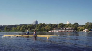 Kürek takımı eğitimi. İki beyaz erkek kürekçinin yan görüntüsü, bir kürek antrenmanı sırasında, Dinyeper nehrinde bir sandalda oturan sporcu, güneşli bir sonbahar günü, sakin bir suda kürek çekiyor. 4k görüntü