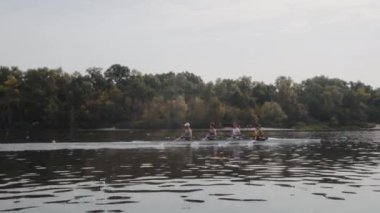 Kürek takımı eğitimi. 4 genç beyaz erkek kürekçinin yan görüntüsü, bir kürek antrenmanı sırasında, Dnipro nehrinde bir sandalda oturan sporcu, sonbaharda sakin bir suda kürek çekiyor. 4K görüntü. Ukrayna, Kyiv 'deki şehir alanı.