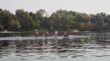 Kürek takımı eğitimi. 4 genç beyaz erkek kürekçinin yan görüntüsü, bir kürek antrenmanı sırasında, Dnipro nehrinde bir sandalda oturan sporcu, sonbaharda sakin bir suda kürek çekiyor. 4K görüntü. Ukrayna, Kyiv 'deki şehir alanı.