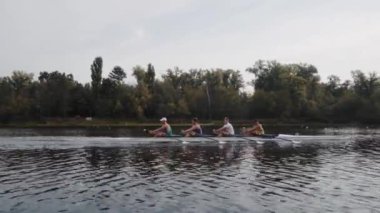 Kürek takımı eğitimi. 4 genç beyaz erkek kürekçinin yan görüntüsü, bir kürek antrenmanı sırasında, Dnipro nehrinde bir sandalda oturan sporcu, sonbaharda sakin bir suda kürek çekiyor. 4K görüntü. Ukrayna, Kyiv 'deki şehir alanı.