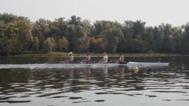 Kürek takımı eğitimi. 4 genç beyaz erkek kürekçinin yan görüntüsü, bir kürek antrenmanı sırasında, Dnipro nehrinde bir sandalda oturan sporcu, sonbaharda sakin bir suda kürek çekiyor. 4K görüntü. Ukrayna, Kyiv 'deki şehir alanı.