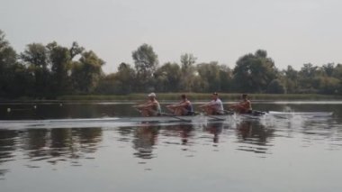 Kürek takımı eğitimi. 4 genç beyaz erkek kürekçinin yan görüntüsü, bir kürek antrenmanı sırasında, Dnipro nehrinde bir sandalda oturan sporcu, sonbaharda sakin bir suda kürek çekiyor. 4K görüntü. Ukrayna, Kyiv 'deki şehir alanı.