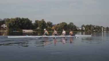 Kürek takımı eğitimi. 4 genç beyaz erkek kürekçinin yan görüntüsü, bir kürek antrenmanı sırasında, Dnipro nehrinde bir sandalda oturan sporcu, sonbaharda sakin bir suda kürek çekiyor. 4K görüntü. Ukrayna, Kyiv 'deki şehir alanı.