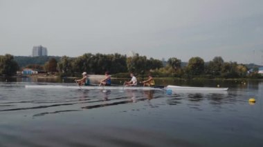 Kürek takımı eğitimi. 4 genç beyaz erkek kürekçinin yan görüntüsü, bir kürek antrenmanı sırasında, Dnipro nehrinde bir sandalda oturan sporcu, sonbaharda sakin bir suda kürek çekiyor. 4K görüntü. Ukrayna, Kyiv 'deki şehir alanı.