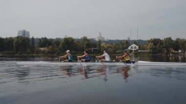 Kürek takımı eğitimi. 4 genç beyaz erkek kürekçinin yan görüntüsü, bir kürek antrenmanı sırasında, Dnipro nehrinde bir sandalda oturan sporcu, sonbaharda sakin bir suda kürek çekiyor. 4K görüntü. Ukrayna, Kyiv 'deki şehir alanı.