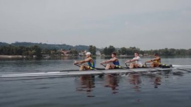 Kürek takımı eğitimi. 4 genç beyaz erkek kürekçinin yan görüntüsü, bir kürek antrenmanı sırasında, Dnipro nehrinde bir sandalda oturan sporcu, sonbaharda sakin bir suda kürek çekiyor. 4K görüntü. Ukrayna, Kyiv 'deki şehir alanı.
