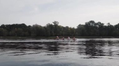 Kürek takımı eğitimi. 4 genç beyaz erkek kürekçinin yan görüntüsü, bir kürek antrenmanı sırasında, Dnipro nehrinde bir sandalda oturan sporcu, sonbaharda sakin bir suda kürek çekiyor. 4K görüntü. Ukrayna, Kyiv 'deki şehir alanı.
