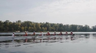 Kürek takımı eğitimi. 8 beyaz erkek kürekçinin yan görüntüsü, bir kürek antrenmanı sırasında, Dinyeper nehrinde bir botta oturan sporcu, sonbaharda sakin bir suda kürek çekiyor. 4K görüntü. Ukrayna, Kyiv 'deki şehir alanı.