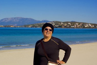 Güneş gözlüklü güzel bir kadın kumsalda kameraya gülümsüyor.