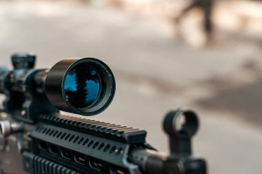 Optik dürbünlü modern bir taktik tüfeğin detaylı görüntüsü. Mavi renkli mercek orman manzarasını yansıtıyor, hassasiyeti ve açık hava kullanımını vurguluyor. Silah kısmi odak noktasında gösteriliyor