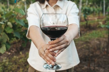Bir kadının iki eli de yemyeşil üzüm bağında büyük bir bardak kırmızı şarap ikram etmesine yakın çekim. Görüntü, misafirperverlik, gelenek ve kırsal çekiciliği çağrıştırıyor.